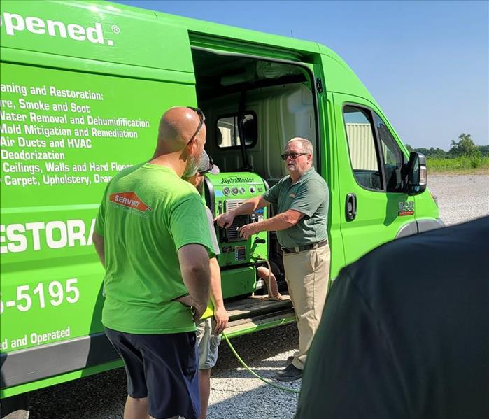 Some employees in green shirts in front of a SERVPRO van with a teacher pointing at a piece of equipment in the van